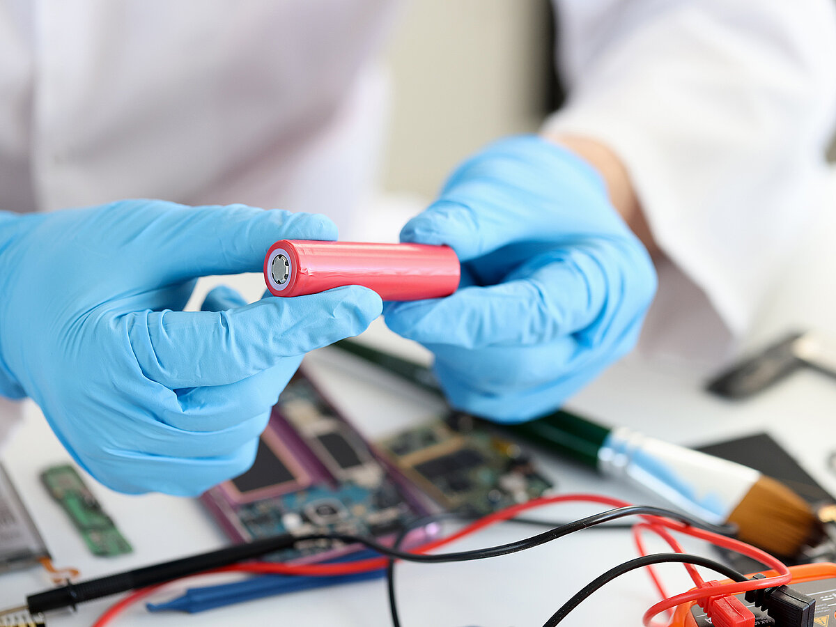 Zwei Hände mit blauen Handschuhen halten eine rote Batterie in einer Werkstattumgebung